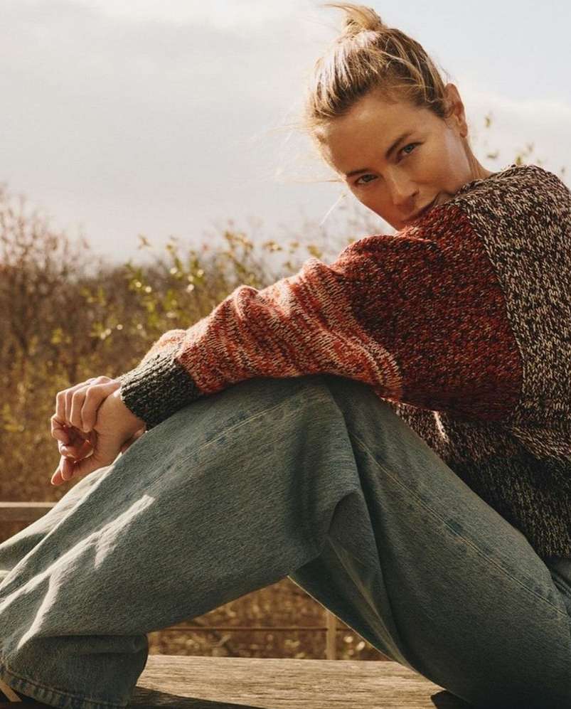 Blonde woman in blue jeans and a colourful sweater sits outdoors with a blurred background of trees and sky in soft, natural light.