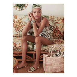 Model seated on a floral print sofa wearing a headscarf, sunglasses, and patterned swimwear with a matching tote bag.