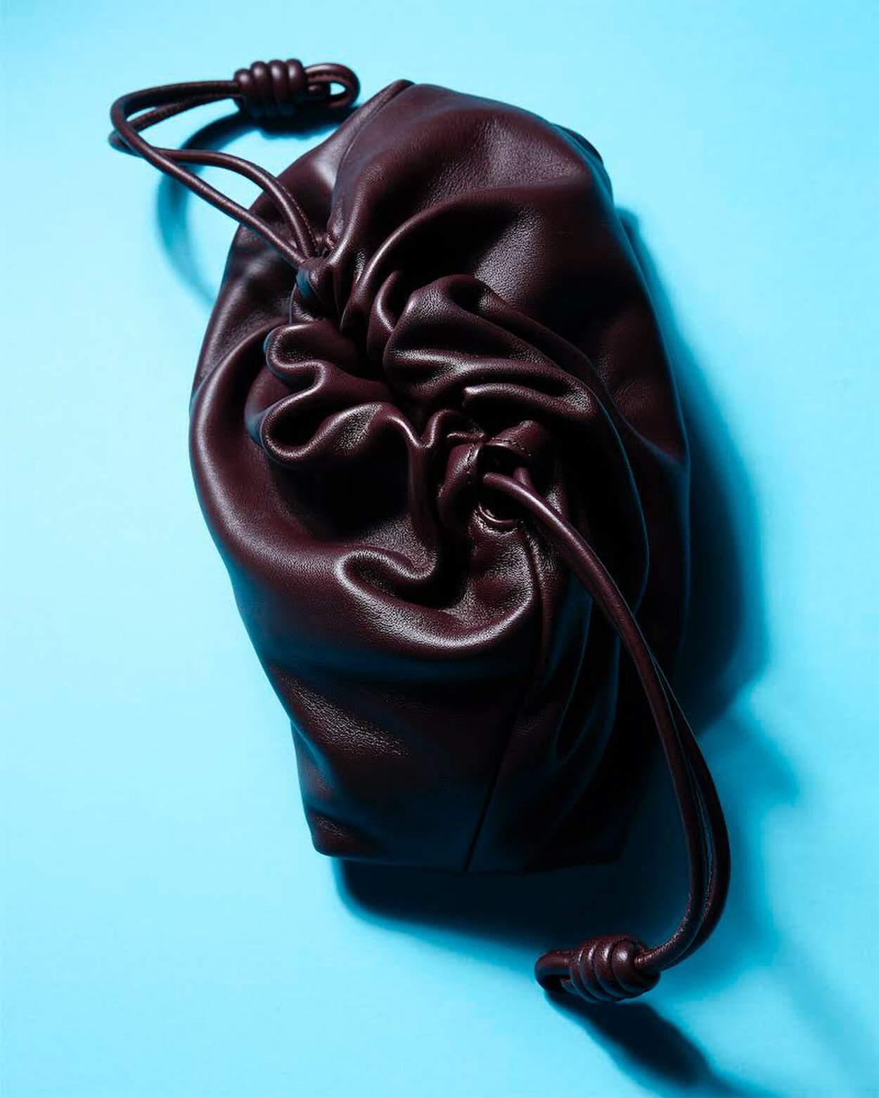 Burgundy-colored leather drawstring pouch bag on a light blue surface, product photography.