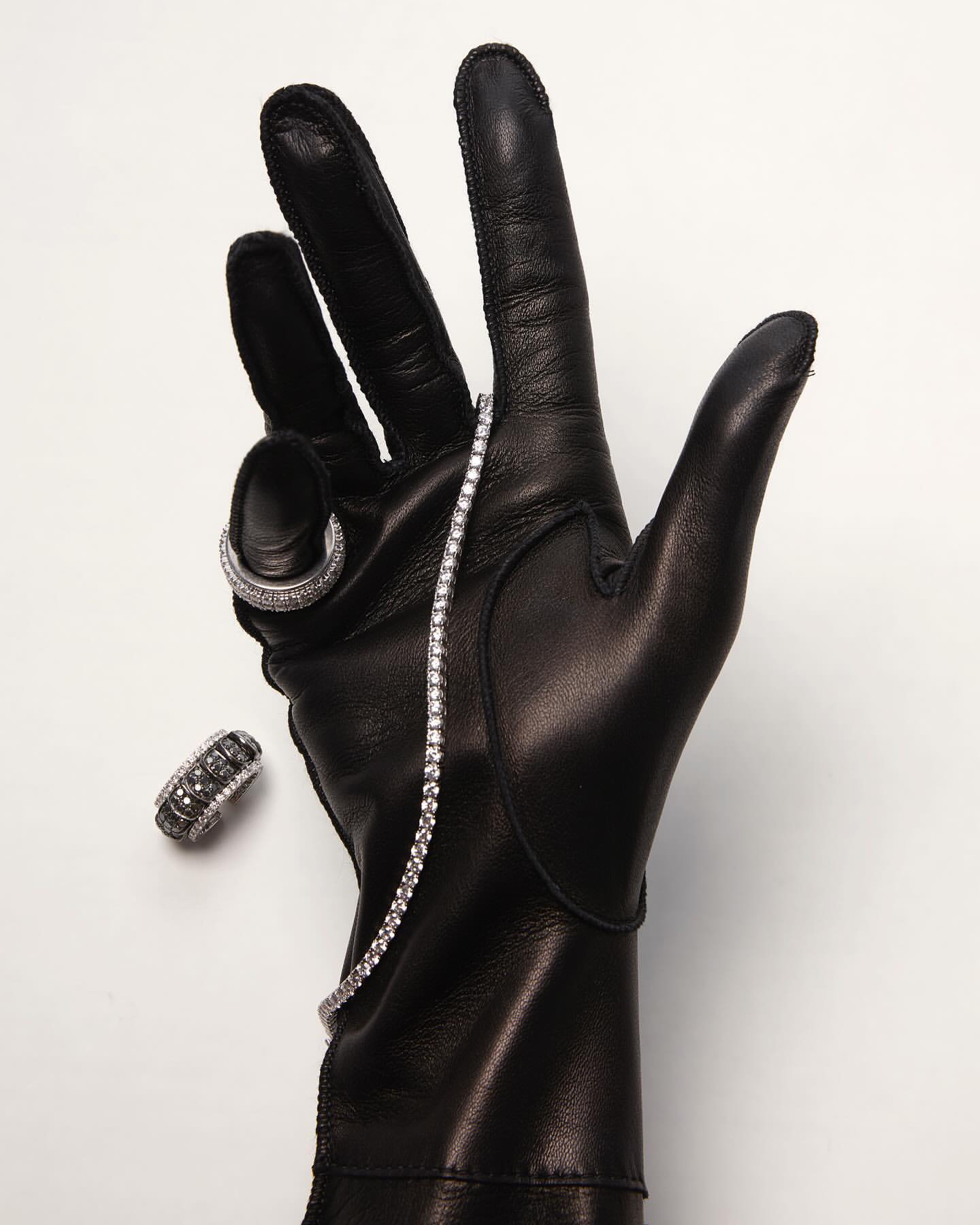 Close-up of a black leather glove displaying diamond jewelry, including a bracelet and rings, against a white backdrop. Studio shot.