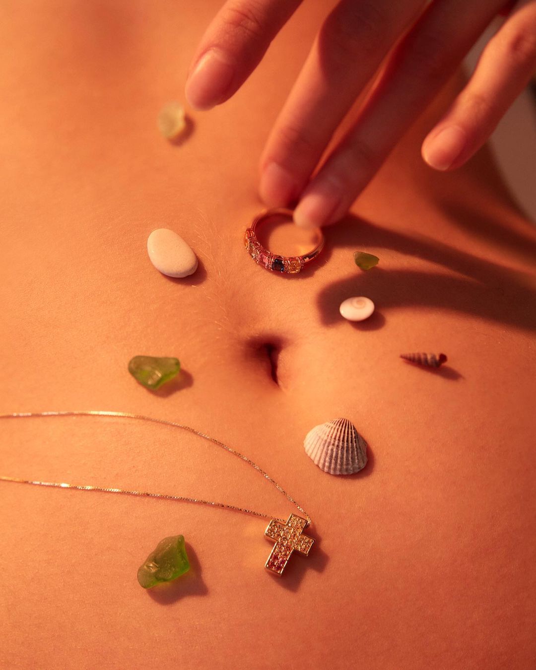 Torso of a person with various small objects and jewelry arranged on the stomach, including shells, pebbles, and a gold necklace.