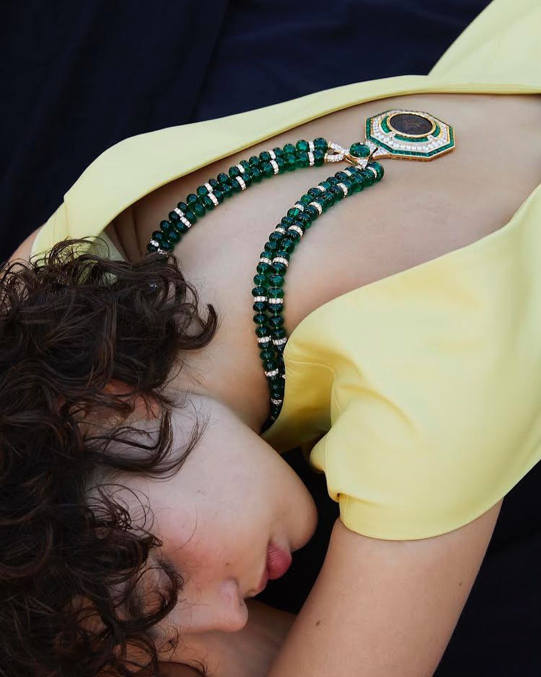 Woman with curly hair poses wearing a yellow blouse and beaded necklaces, against a dark backdrop, jewelry fashion advertisement.