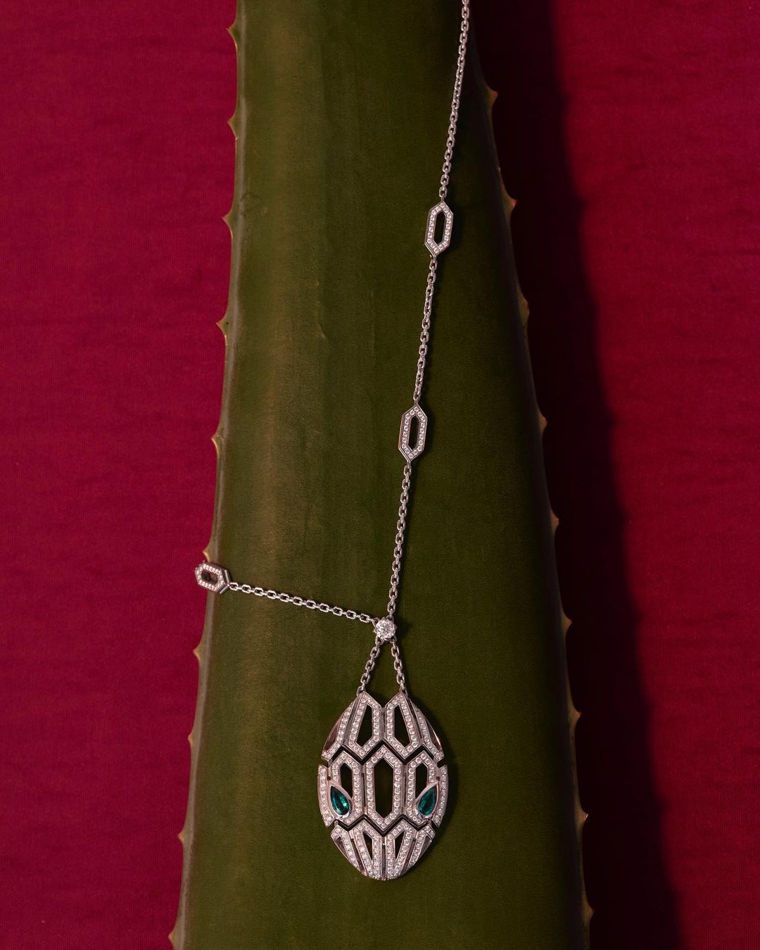 Silver chain necklace featuring geometric shapes and teardrop pendant with green stones, lying on a green aloe leaf against red backdrop.