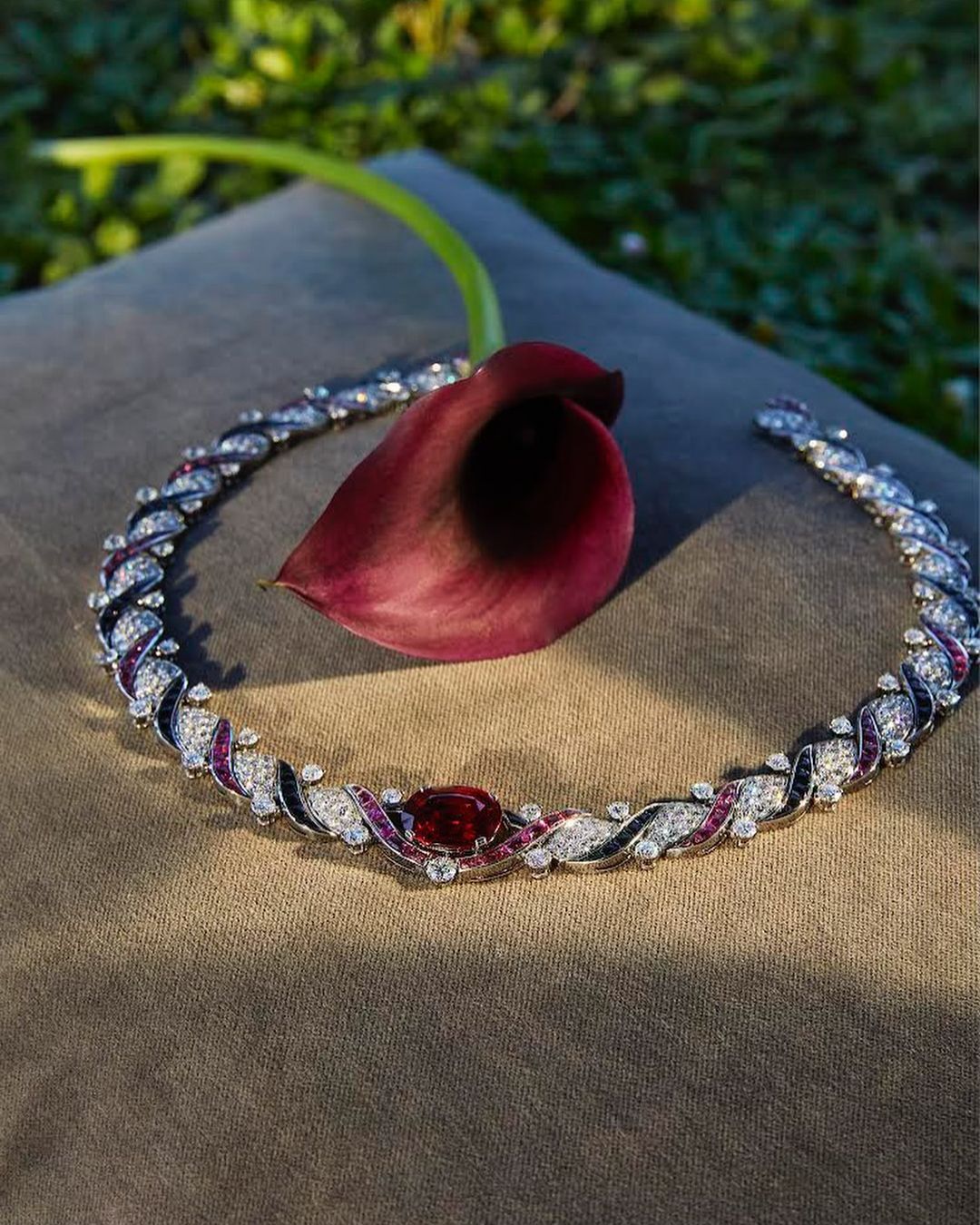 Elegant diamond collar necklace with central ruby and dark calla lily flower on a gray cushion against a green grass background.