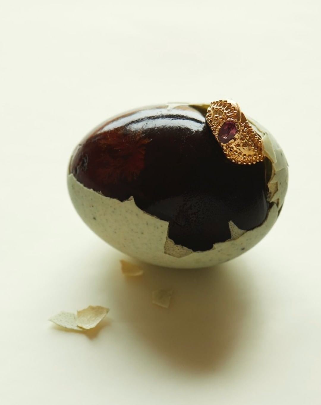 Close-up shot of a century egg with cracked shell, topped with an ornate gold ring featuring a red stone, on a white surface.