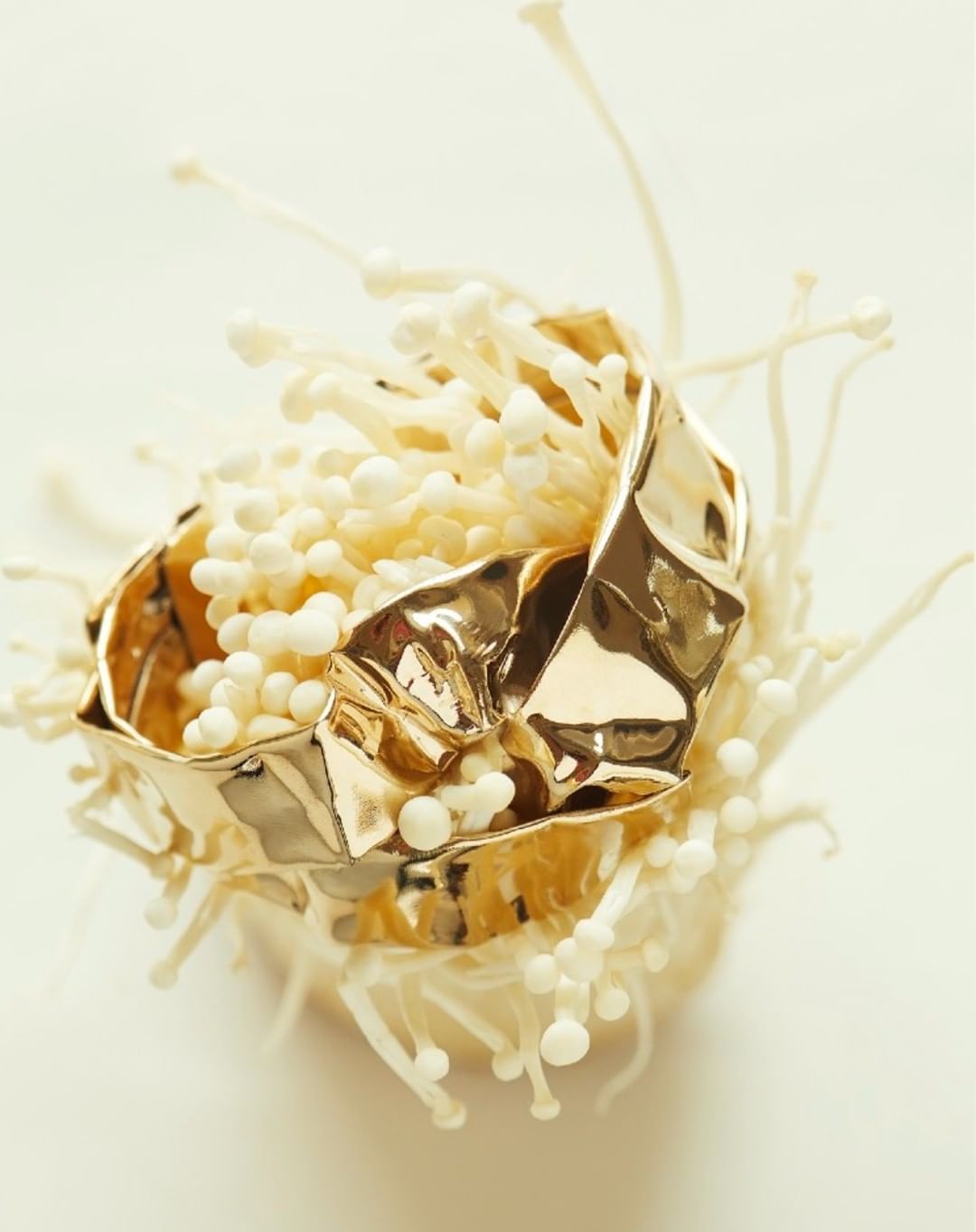 Close-up of a shiny gold cuff bracelet displayed in a nest of delicate, white enoki mushrooms on an off-white surface.