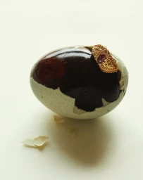 An ornate gold ring with a pink gemstone rests on a cracked dark speckled egg against a pale background.