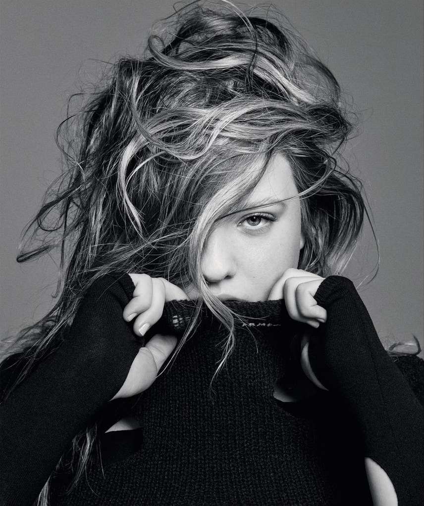 Black and white portrait of a young woman with wavy hair partially covering her face, wearing a black sweater pulled up to her chin.