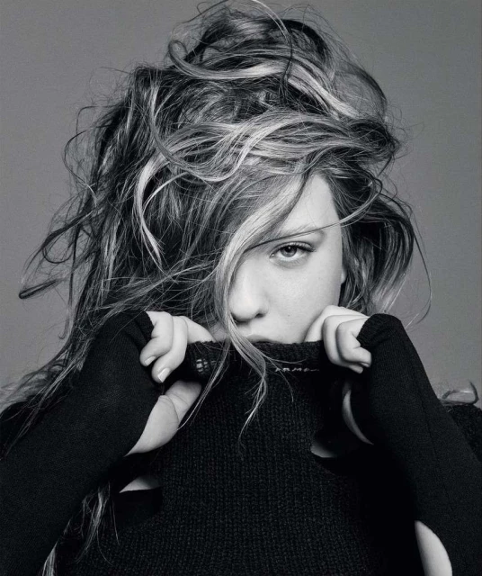 Black and white portrait of a young woman with wavy hair partially covering her face, wearing a black sweater pulled up to her chin.