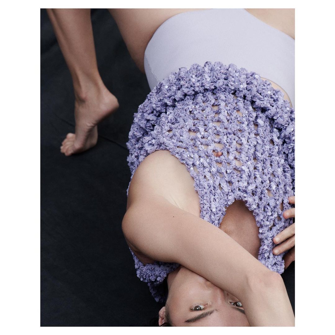 Upside-down view of a person in lilac top and bottoms against a dark background, with only the lower legs visible at the top.