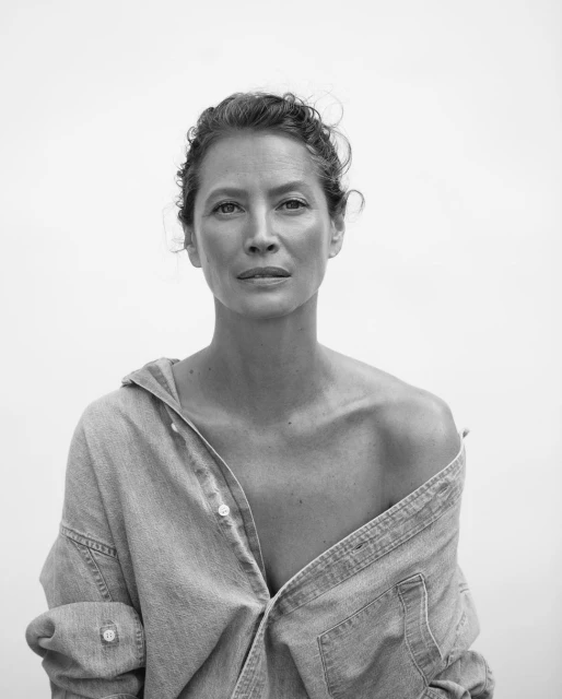 Black and white portrait of a mature woman with an open denim shirt, casually draped off her shoulders, against a neutral background.