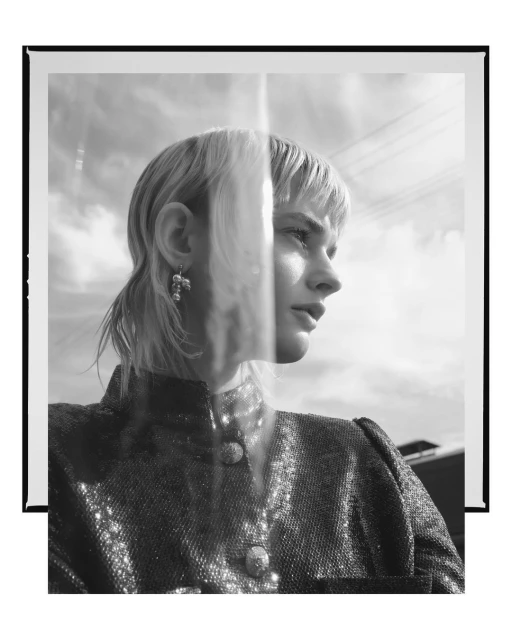 Black and white portrait of a blonde woman in a sparkling jacket, posing indoors in front of a frame with a sky background.