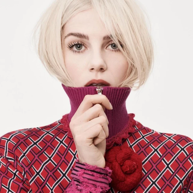 Close-up portrait of a blonde woman with red lipstick wearing a patterned red and white jacket with a high magenta collar.