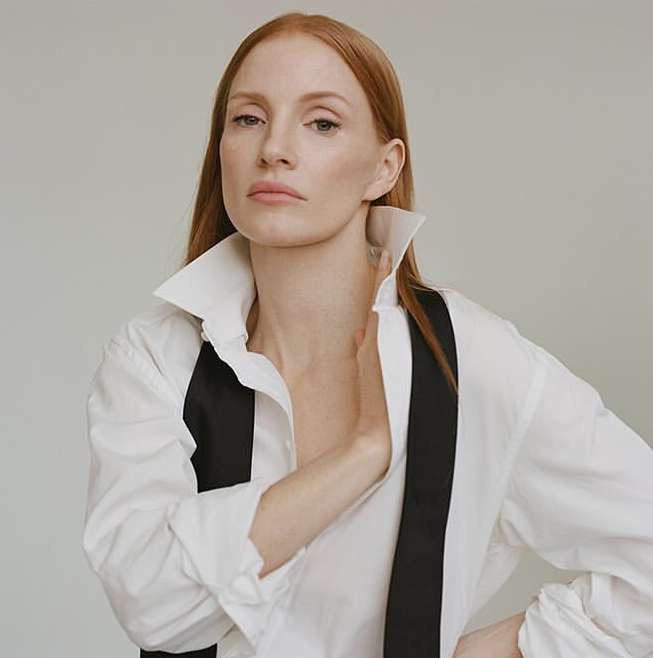 A portrait of a red-haired woman wearing a white shirt and black suspenders against a neutral background in a studio setting.
