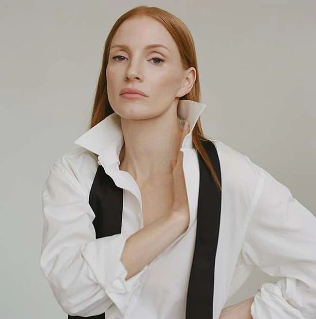A portrait of a red-haired woman wearing a white shirt and black suspenders against a neutral background in a studio setting.