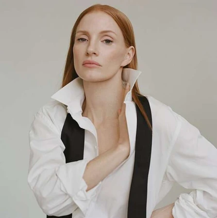 A portrait of a red-haired woman wearing a white shirt and black suspenders against a neutral background in a studio setting.