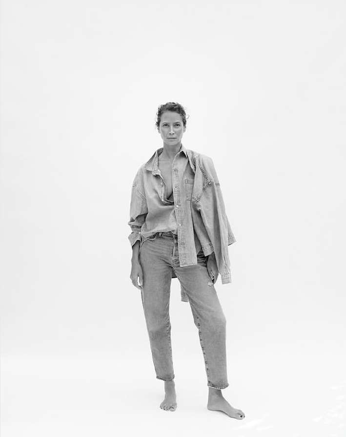 Monochrome portrait of a woman in an unbuttoned denim shirt and jeans against a bright white backdrop in a full length shot.
