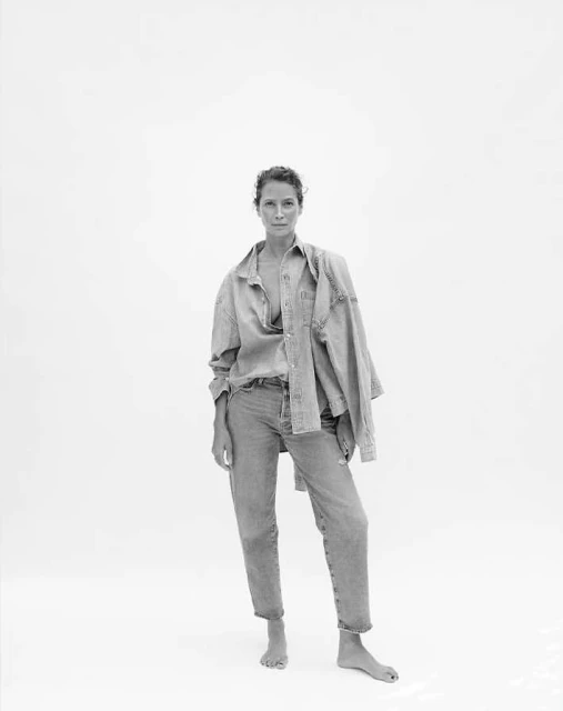 Monochrome portrait of a woman in an unbuttoned denim shirt and jeans against a bright white backdrop in a full length shot.