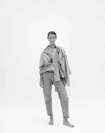 Monochrome portrait of a woman in an unbuttoned denim shirt and jeans against a bright white backdrop in a full length shot.