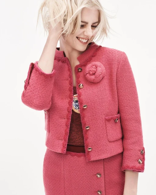 A blonde woman in a pink tweed Chanel jacket and skirt smiles and glances down against a bright white background in a studio shot.