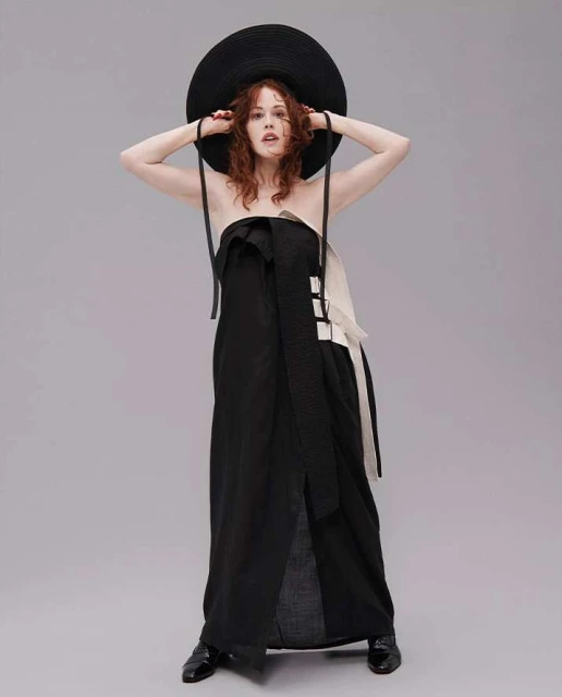 An adult female model wearing a black, strapless dress and a black hat, with her hands touching the hat, stands in a studio setting.