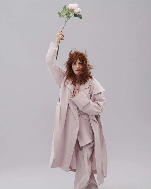 A woman with red hair, in a pale pink pantsuit and coat, holds a single pink flower against a light gray background in a studio setting.