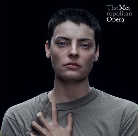 Close-up studio portrait of an adult female with short hair wearing a gray crewneck shirt, posing in front of a black backdrop.