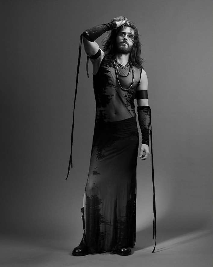 Full-length studio portrait of a bearded adult male model in a sheer top and long skirt, against a grey backdrop in black and white.