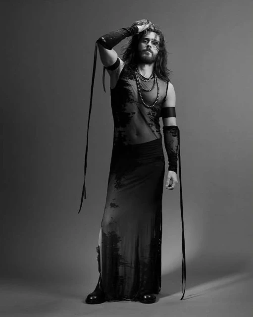 Full-length studio portrait of a bearded adult male model in a sheer top and long skirt, against a grey backdrop in black and white.