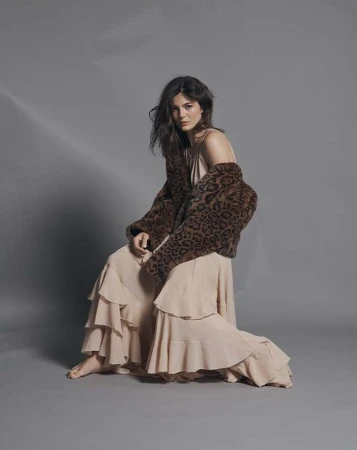 A brunette woman poses in a beige dress and leopard print jacket against a grey backdrop, showcasing a blend of fashion and natural beauty.