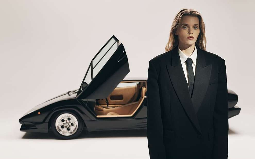 Woman in black suit poses with black Lamborghini car, open doors, studio background, fashion and automotive editorial style.
