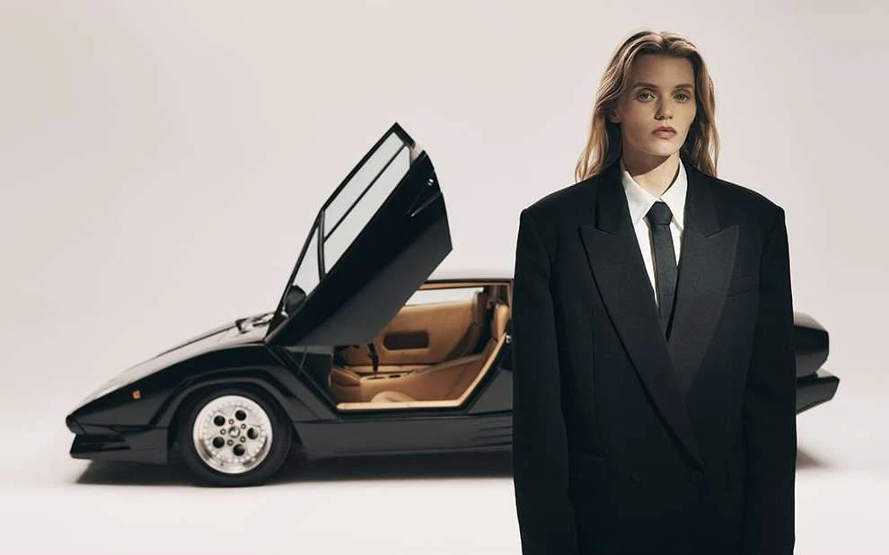 Woman in black suit poses with black Lamborghini car, open doors, studio background, fashion and automotive editorial style.