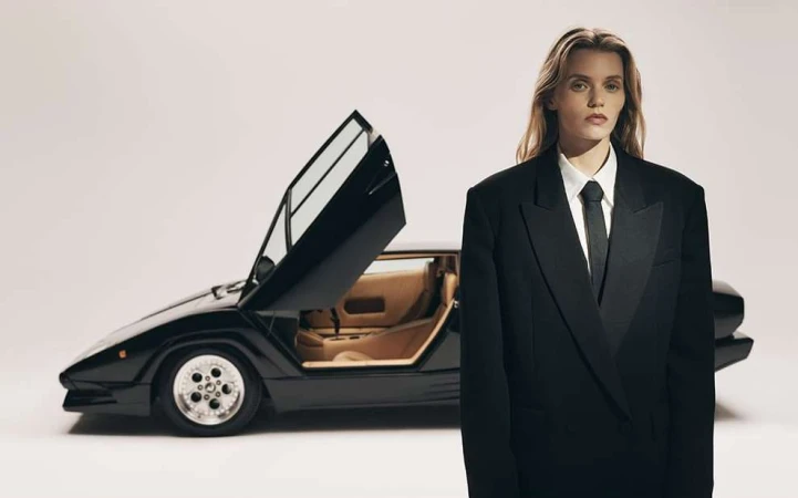 Woman in black suit poses with black Lamborghini car, open doors, studio background, fashion and automotive editorial style.