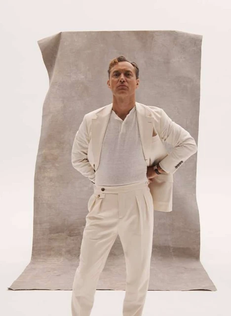 Portrait of a man in a cream-colored suit posing in front of a textured backdrop. He is fair-skinned with light brown hair.