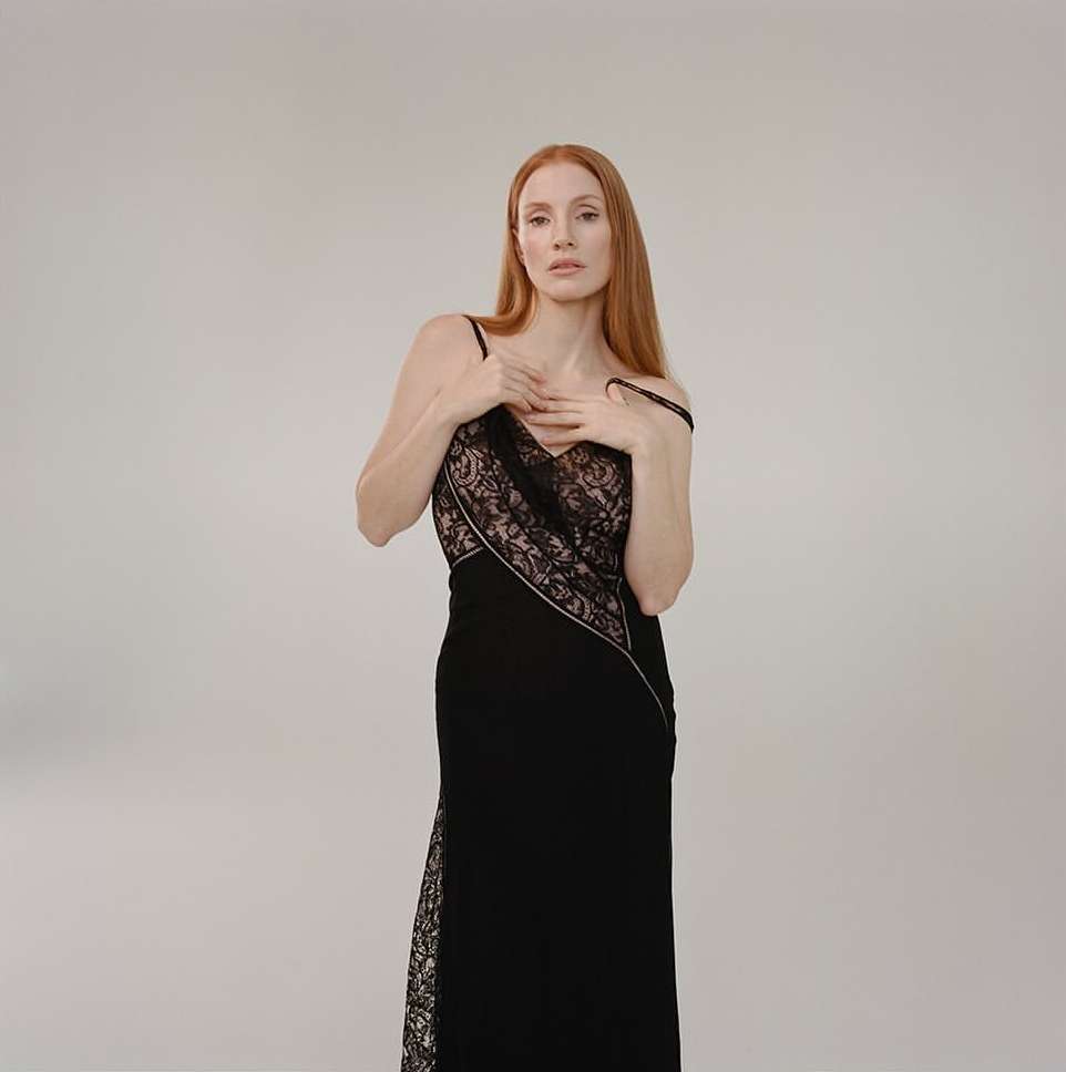 Elegant woman with long red hair poses in a black formal gown with lace detail in a photo studio setting against a neutral backdrop.