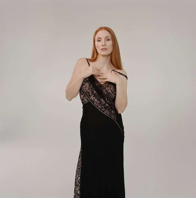 Elegant woman with long red hair poses in a black formal gown with lace detail in a photo studio setting against a neutral backdrop.