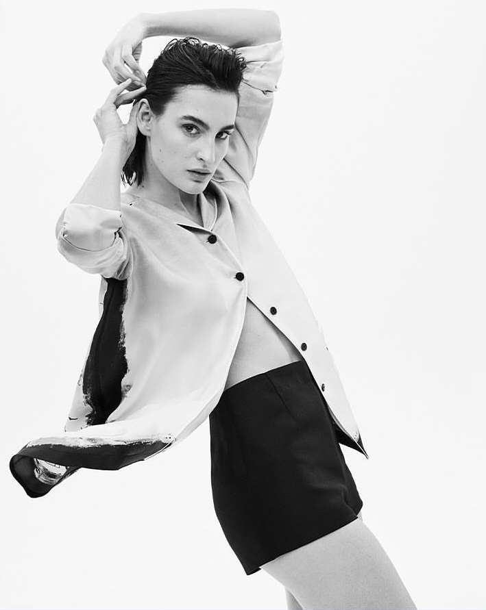 Monochrome portrait of a fair-skinned adult woman in short shorts and an unbuttoned jacket posing against a white background.