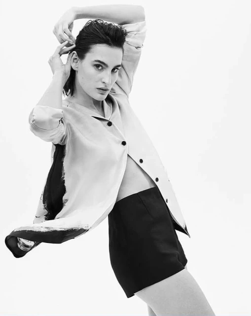 Monochrome portrait of a fair-skinned adult woman in short shorts and an unbuttoned jacket posing against a white background.