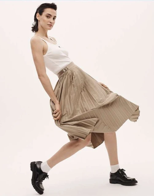 A woman poses in a pleated skirt, a tank top, and black shoes against a white backdrop, showcasing a minimalist fashion aesthetic.
