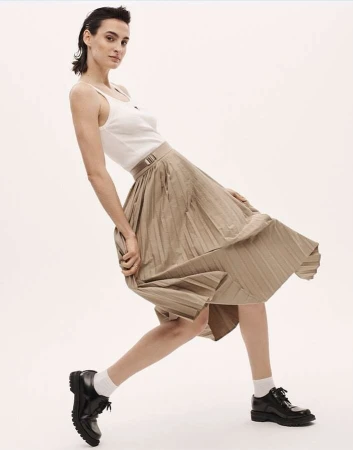 A woman poses in a pleated skirt, a tank top, and black shoes against a white backdrop, showcasing a minimalist fashion aesthetic.