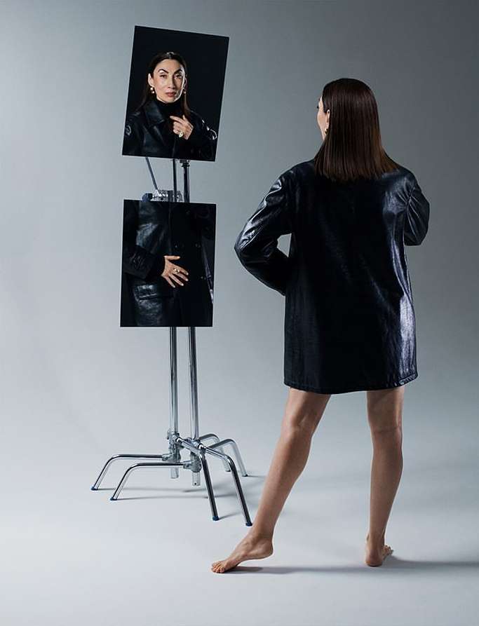 An adult woman in a black jacket stands barefoot in front of mirrors showing her reflection in a fashion shot against a grey background.