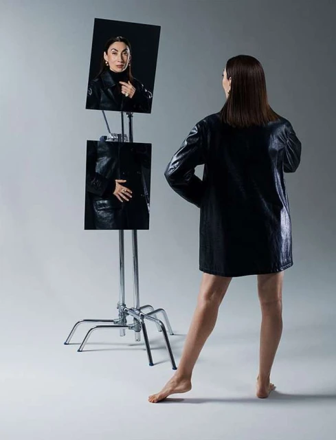 An adult woman in a black jacket stands barefoot in front of mirrors showing her reflection in a fashion shot against a grey background.