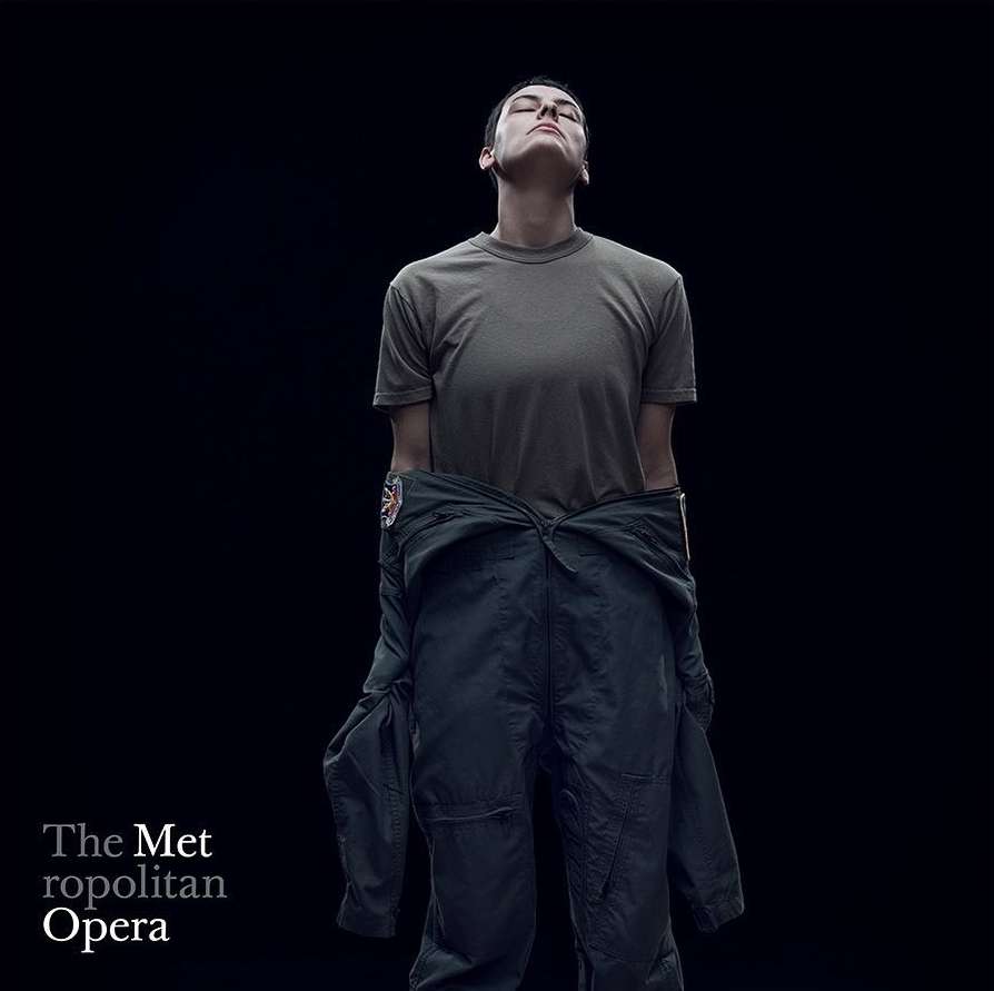 Adult with closed eyes in olive T-shirt and navy flight suit stands in a photo studio with dark background for Metropolitan Opera.