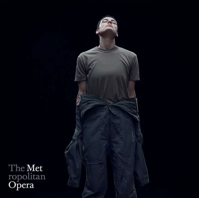 Adult with closed eyes in olive T-shirt and navy flight suit stands in a photo studio with dark background for Metropolitan Opera.