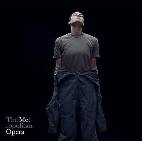 Adult with closed eyes in olive T-shirt and navy flight suit stands in a photo studio with dark background for Metropolitan Opera.