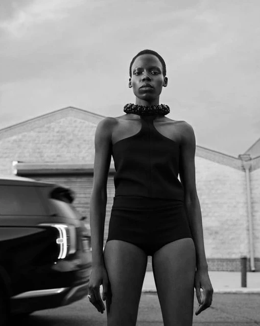 Monochrome portrait of a woman in a black top and shorts, standing outdoors with a car and building in the background, fashion.