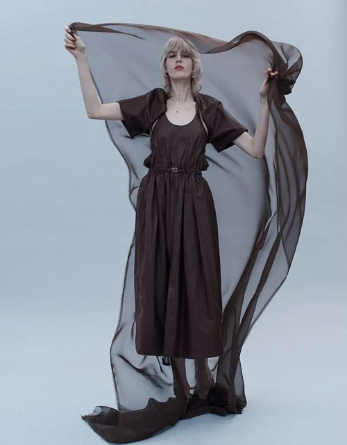 A woman with fair skin and short blonde hair poses in a brown dress, holding a sheer brown fabric in a full shot against a grey background.