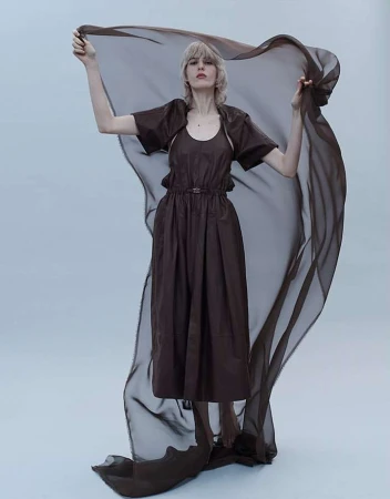 A woman with fair skin and short blonde hair poses in a brown dress, holding a sheer brown fabric in a full shot against a grey background.