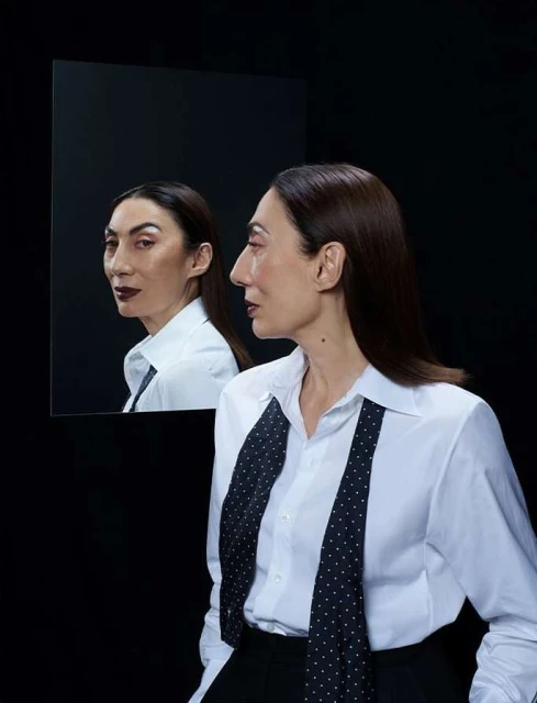 Stylish portrait of a mature adult woman with dark hair wearing a white button-down shirt and tie, standing in front of a mirror.