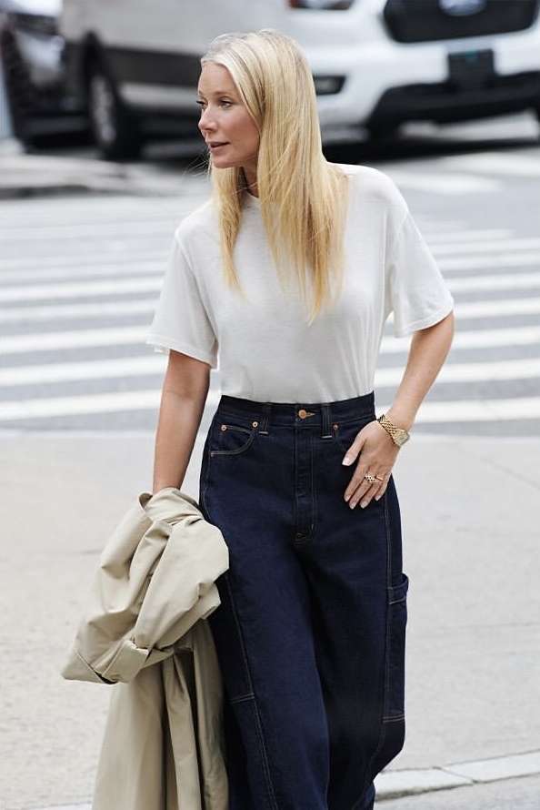 Gwyneth Paltrow wearing a white t-shirt and dark jeans holding a trench coat on a city street.